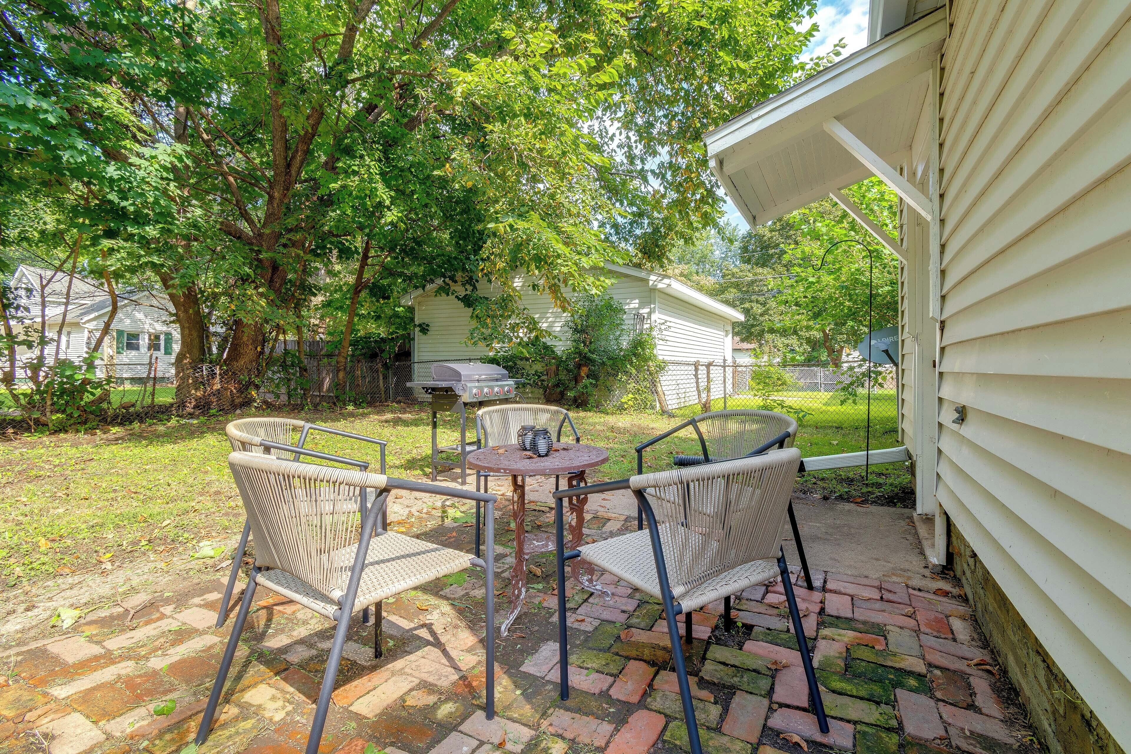 A patio with wicker chairs and a table, surrounded by a brick pathway and greenery.