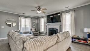 Ein Wohnzimmer mit einem großen beigefarbenen Sofa, einem Kamin, einem Deckenventilator und einem Flachbildfernseher, der über dem Kamin angebracht ist.