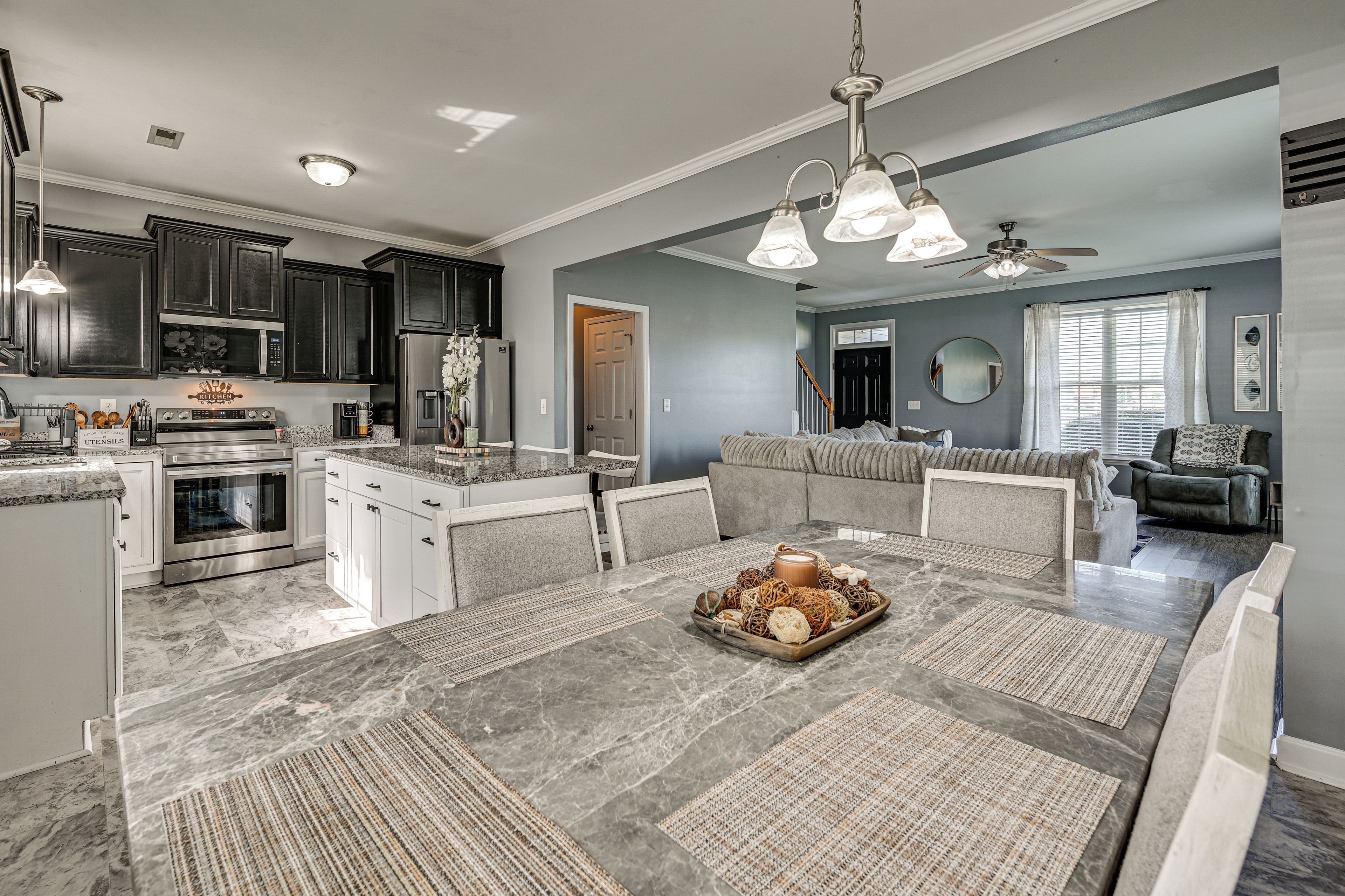 A modern kitchen with dark cabinets, a dining area with a marble table, and a living room with a gray sofa.