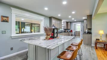A kitchen with a marble island, wooden bar stools, and a view of the backyard through a large window.