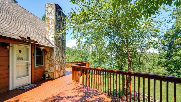 A wooden deck with a stone chimney, a door with a welcome sign, and a view of a forested area. - Pinnacle Lakefront Retreat: Dock, Canoe & Fire Pit (Pinnacle)