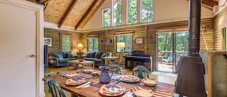 A log cabin interior with a dining area, sofas, a piano, and a wood stove.