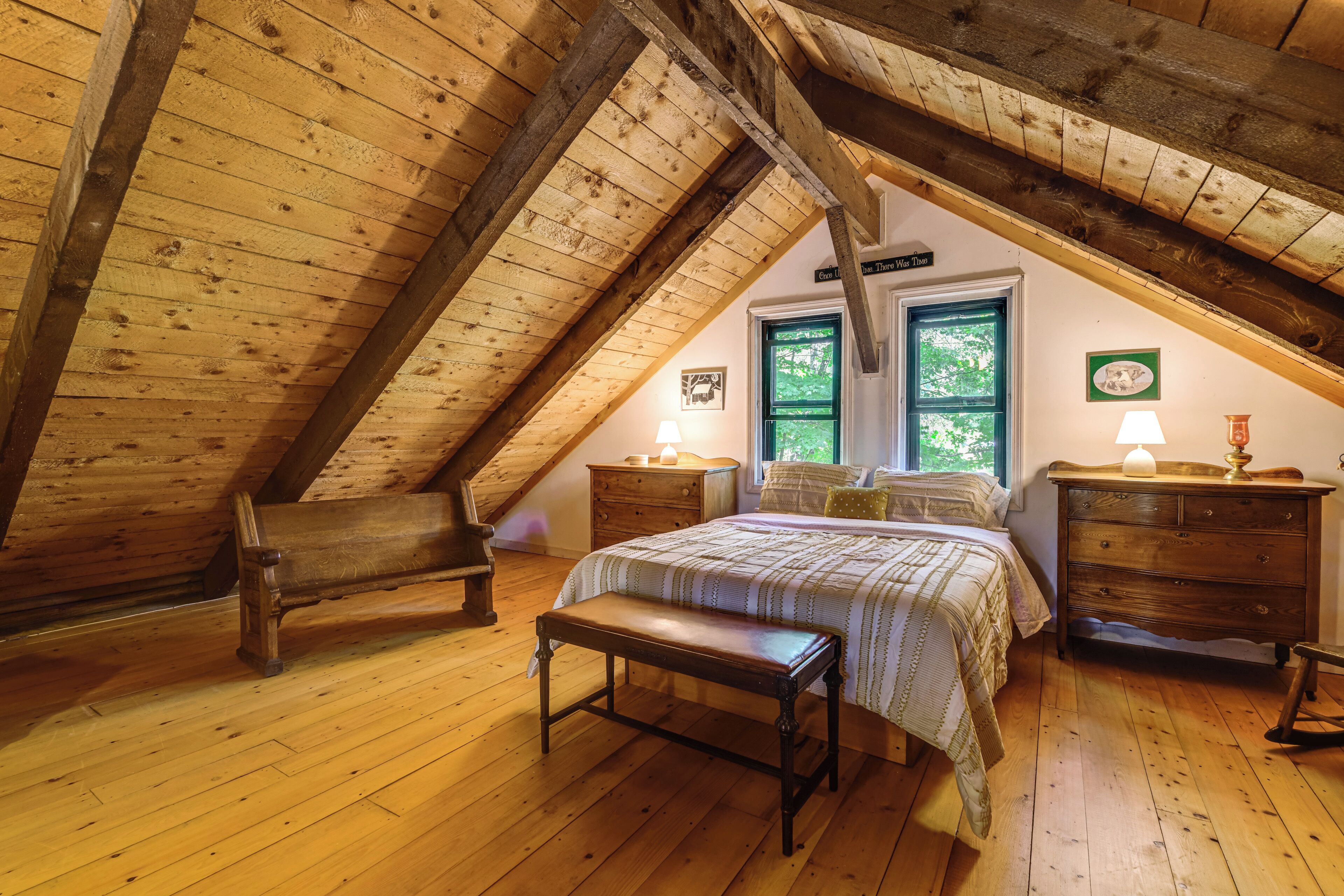 A cozy attic bedroom with a sloped ceiling, wooden floors, a bed with a quilt, two nightstands, a bench, and a dresser.