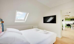 Habitación de hotel moderna con cama, televisor de pantalla plana, tragaluz y vista al área de la cocina.