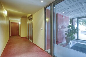 Un couloir avec un sol recouvert de moquette, une porte au fond et une plante en pot près d’une porte vitrée.
