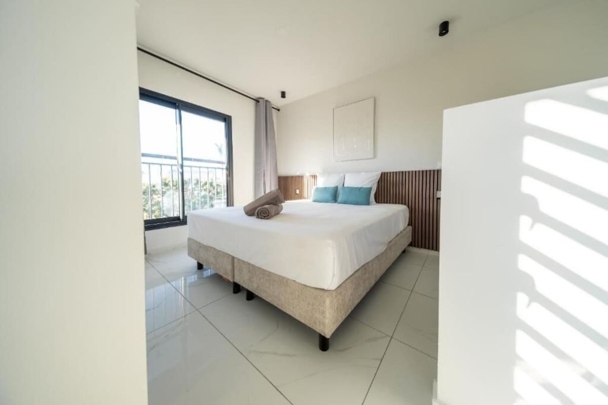 Une chambre moderne avec un grand lit, une vue sur l’extérieur par une fenêtre et un luminaire au plafond.
