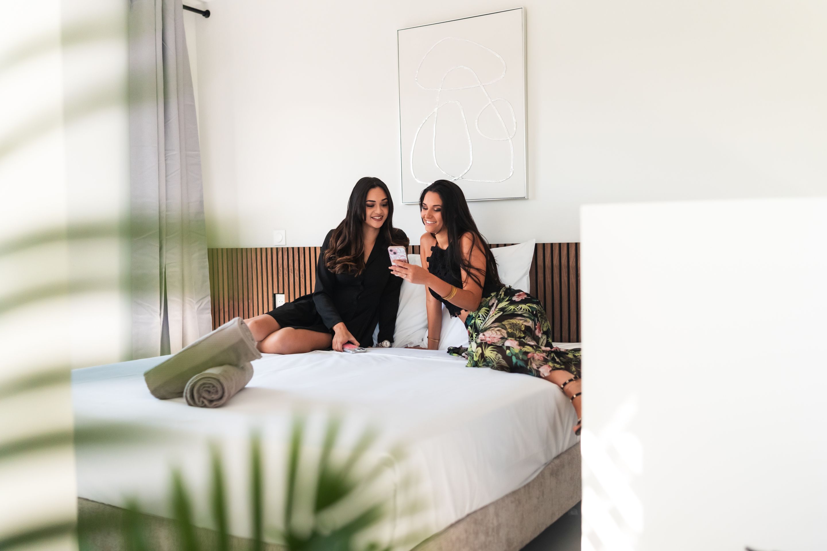Deux femmes sont assises sur un lit dans une chambre d’hôtel, l’une d’elles tient un téléphone.