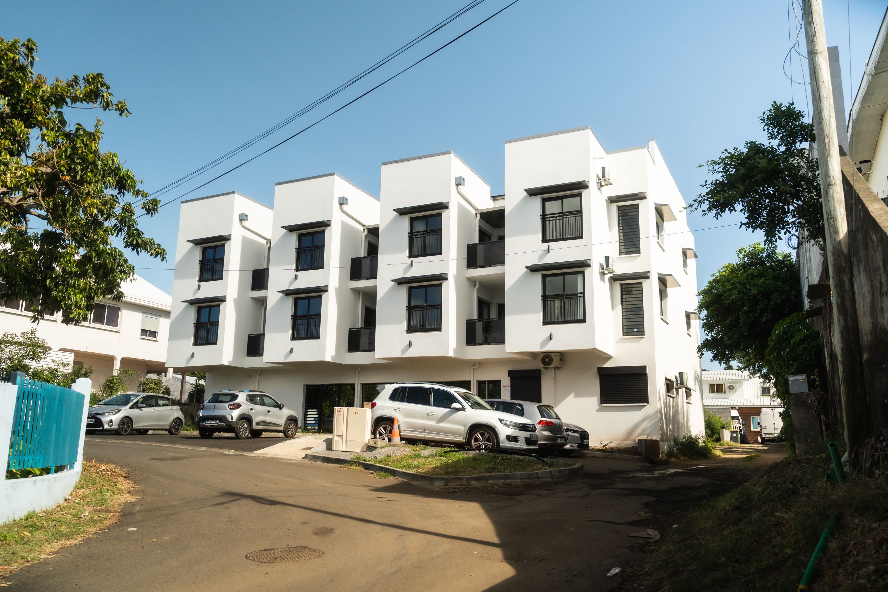 A modern multi-story residential building with a flat roof, multiple balconies, and parked cars in the driveway.