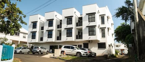 Un moderno edificio residencial de varios pisos con techo plano, varios balcones y coches aparcados en la entrada.