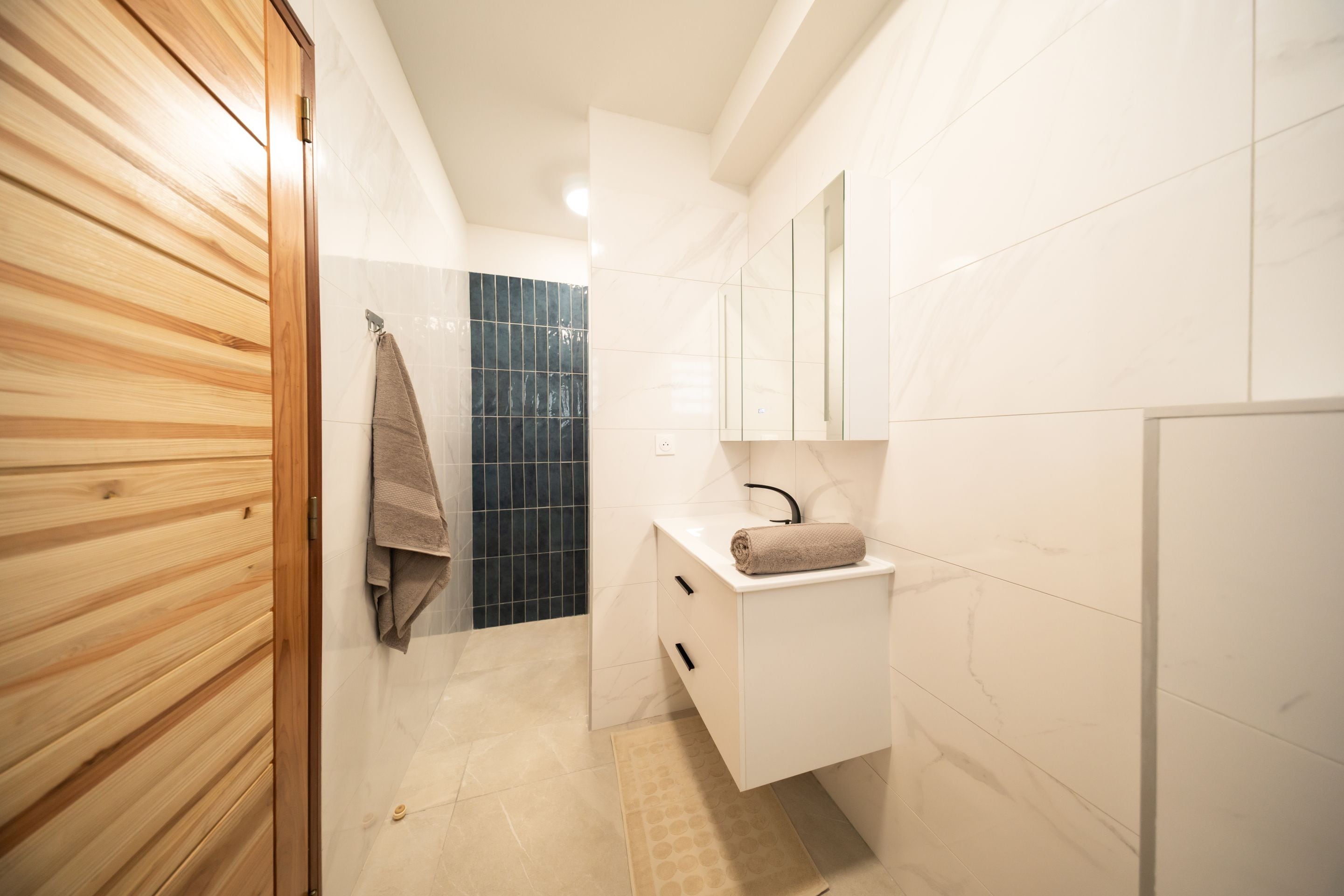 A modern bathroom with a wooden door, a towel hanging on the wall, a sink with a mirror, and tiled walls.