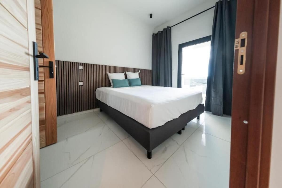 Une chambre d’hôtel avec un lit, une tête de lit en bois, une fenêtre avec des rideaux et une porte avec une poignée.