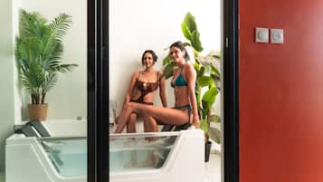 Dos personas sentadas en una bañera con una planta en maceta y una pared roja al fondo.