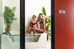 Dos personas sentadas en una bañera con una planta en maceta y una pared roja al fondo.