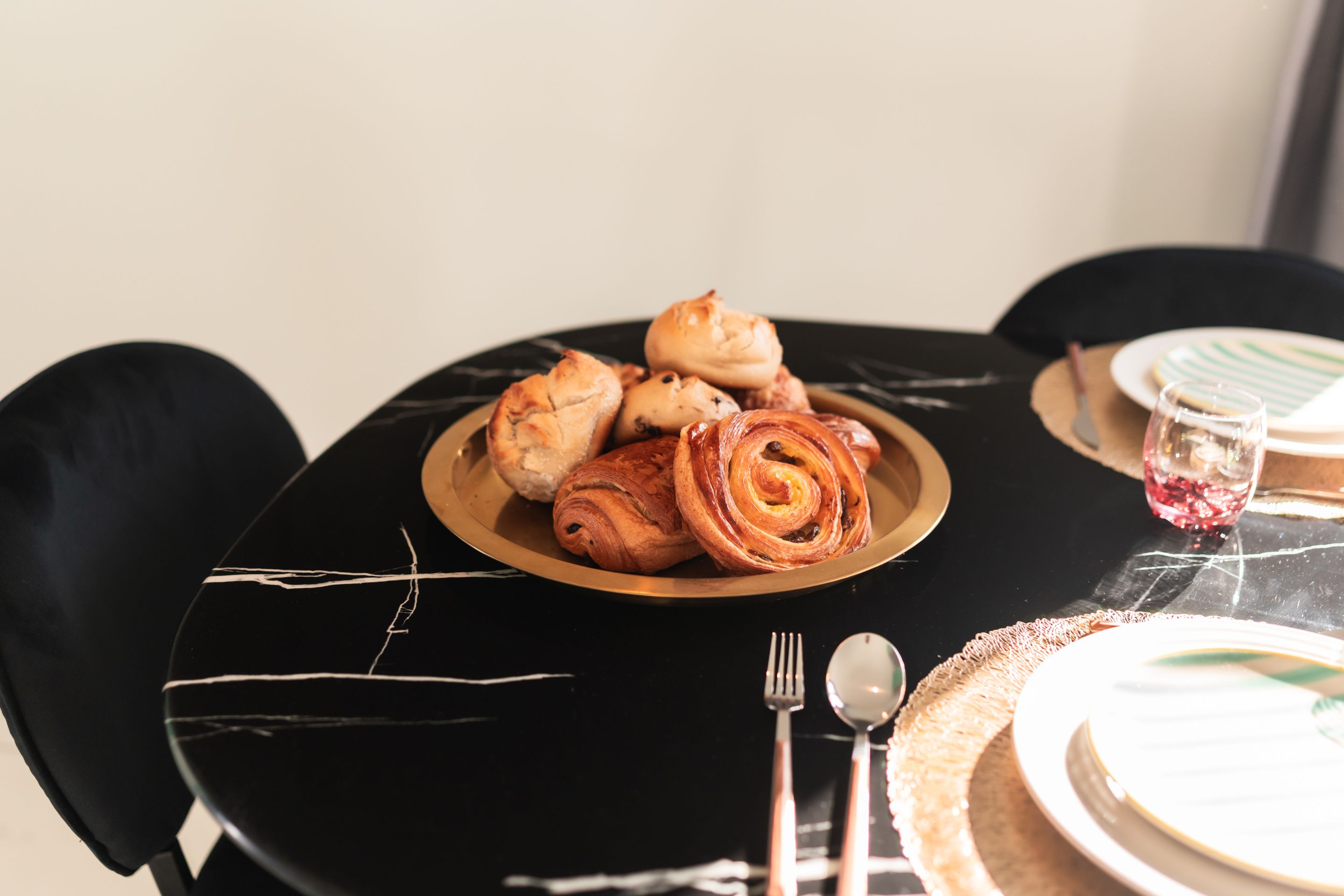 A dining table with a plate of pastries, cutlery, and a glass of red beverage.