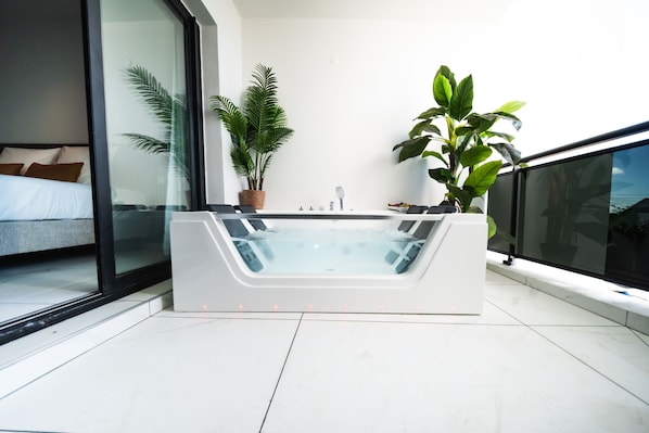 Un baño moderno con bañera de hidromasaje con cerramiento de vidrio, una planta grande en maceta y vistas al dormitorio a través de una puerta de cristal.