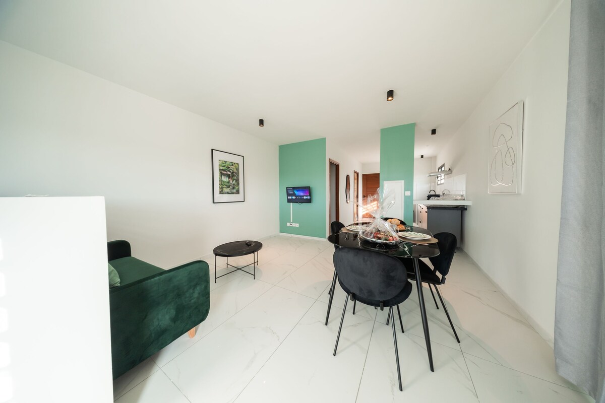 Una sala de estar moderna con un sofá verde, una mesa de comedor con sillas, un televisor montado en la pared y una cocina al fondo.