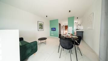 A modern living room with a green sofa, a dining table with chairs, a TV mounted on the wall, and a kitchen area in the background.