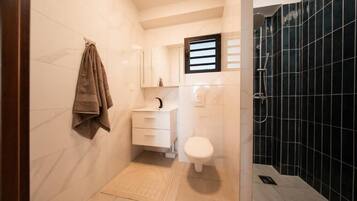 A compact bathroom with a white sink, a white toilet, a towel rack, and a shower area with dark tiles.