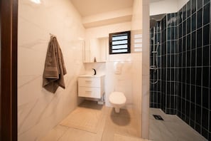 Une petite salle de bain avec un lavabo blanc, des toilettes blanches, un porte-serviettes et un espace douche aux carreaux foncés.