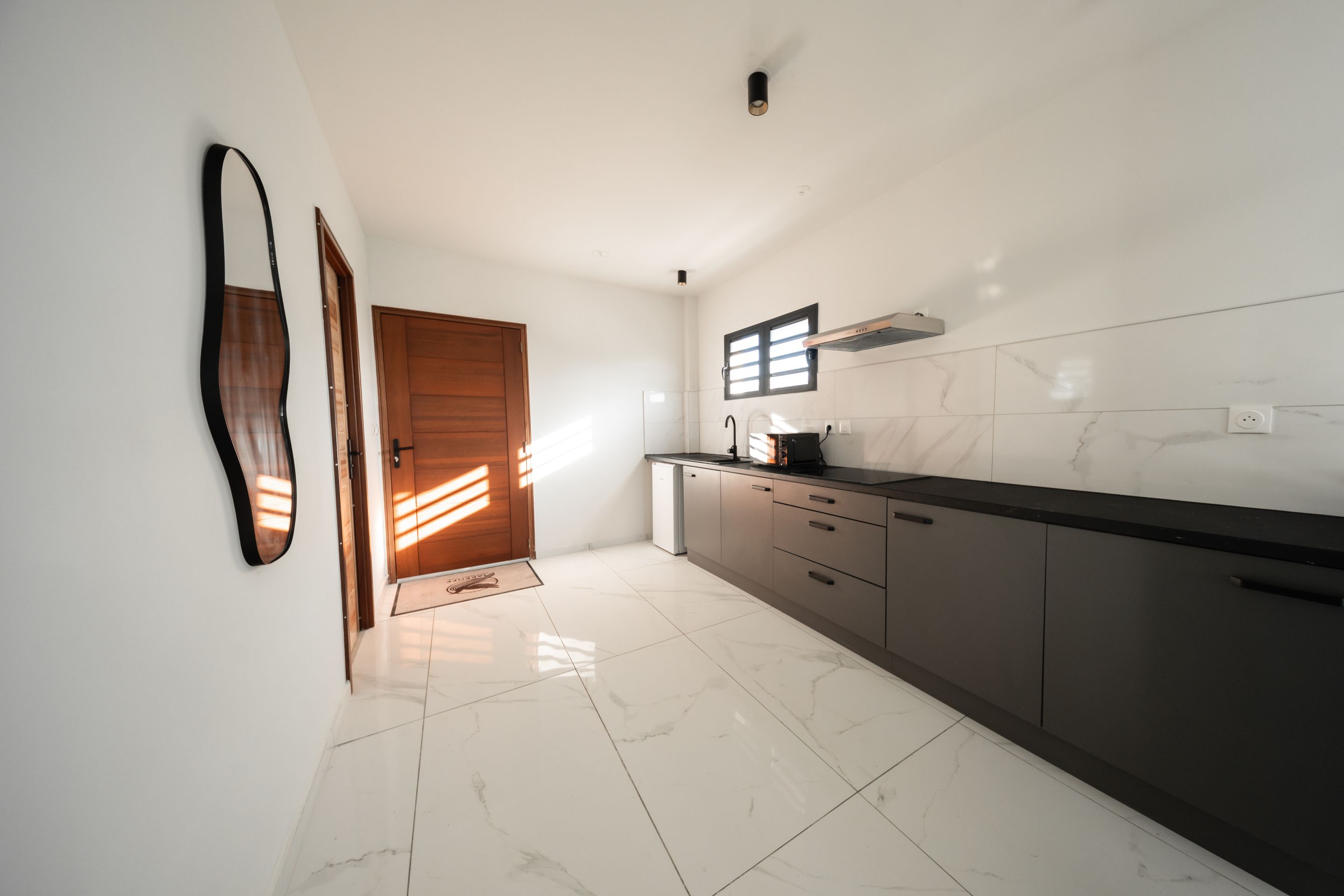 A modern kitchen with white marble tiles, dark cabinetry, and a wooden door.