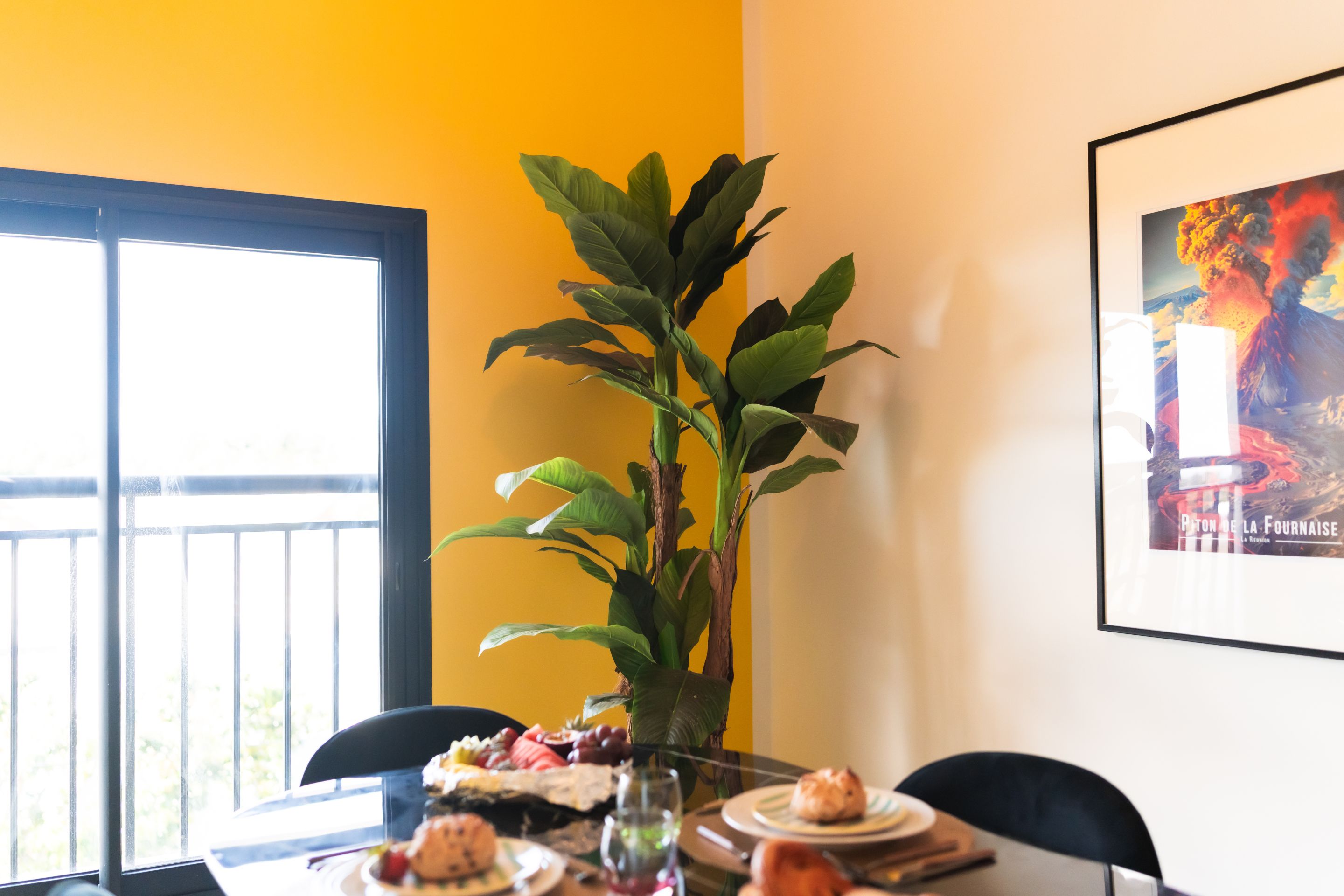 Un coin repas avec une table dressée pour deux personnes, une plante en pot et un tableau encadré au mur.