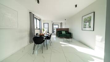 A modern living room with a dining area, a green sofa, and a large window providing natural light.