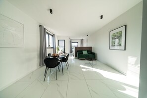 A modern living room with a dining area, a green sofa, and a large window providing natural light.