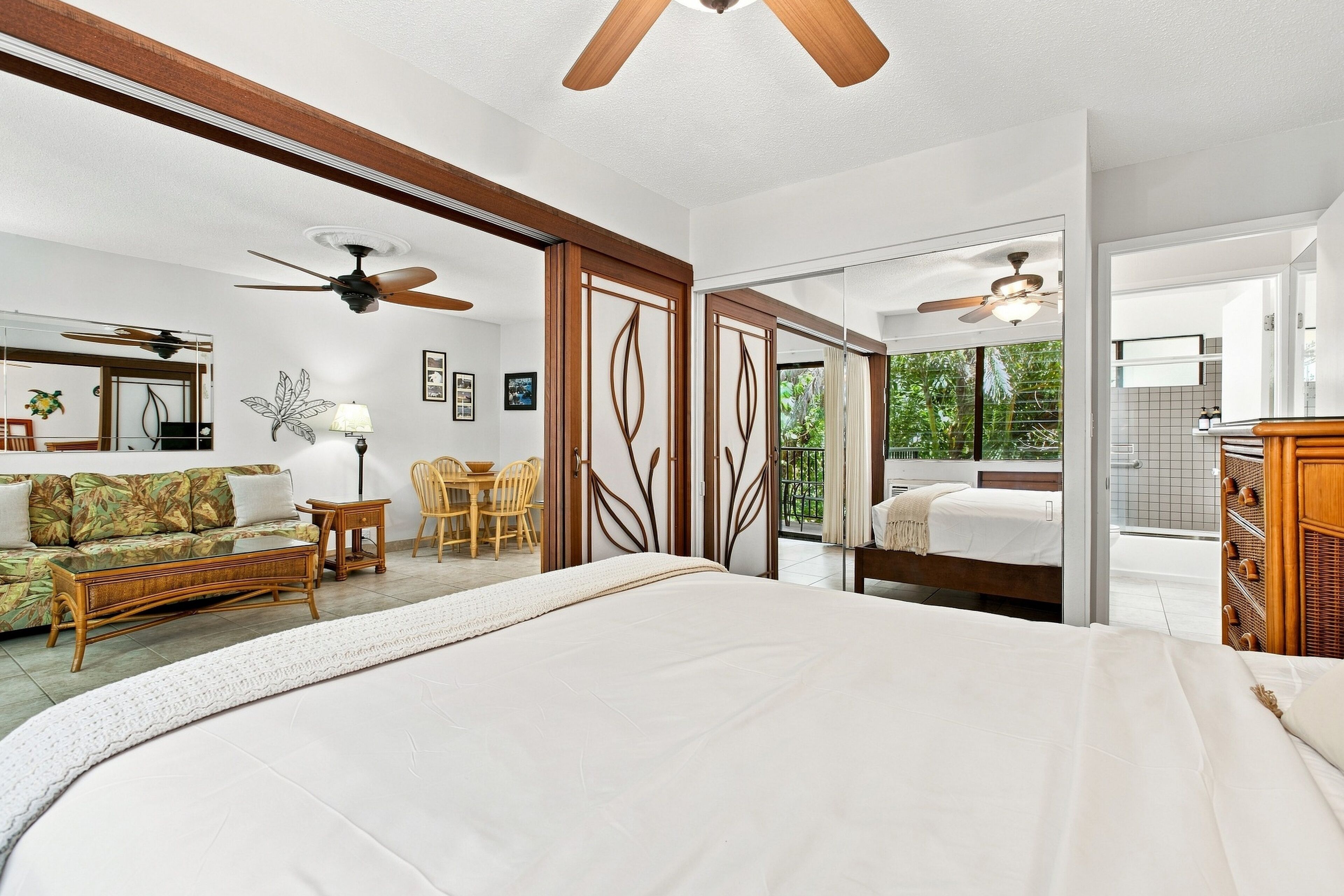 A bedroom with a large bed, a ceiling fan, a dining area with chairs and a table, and a view through a sliding door to a balcony.