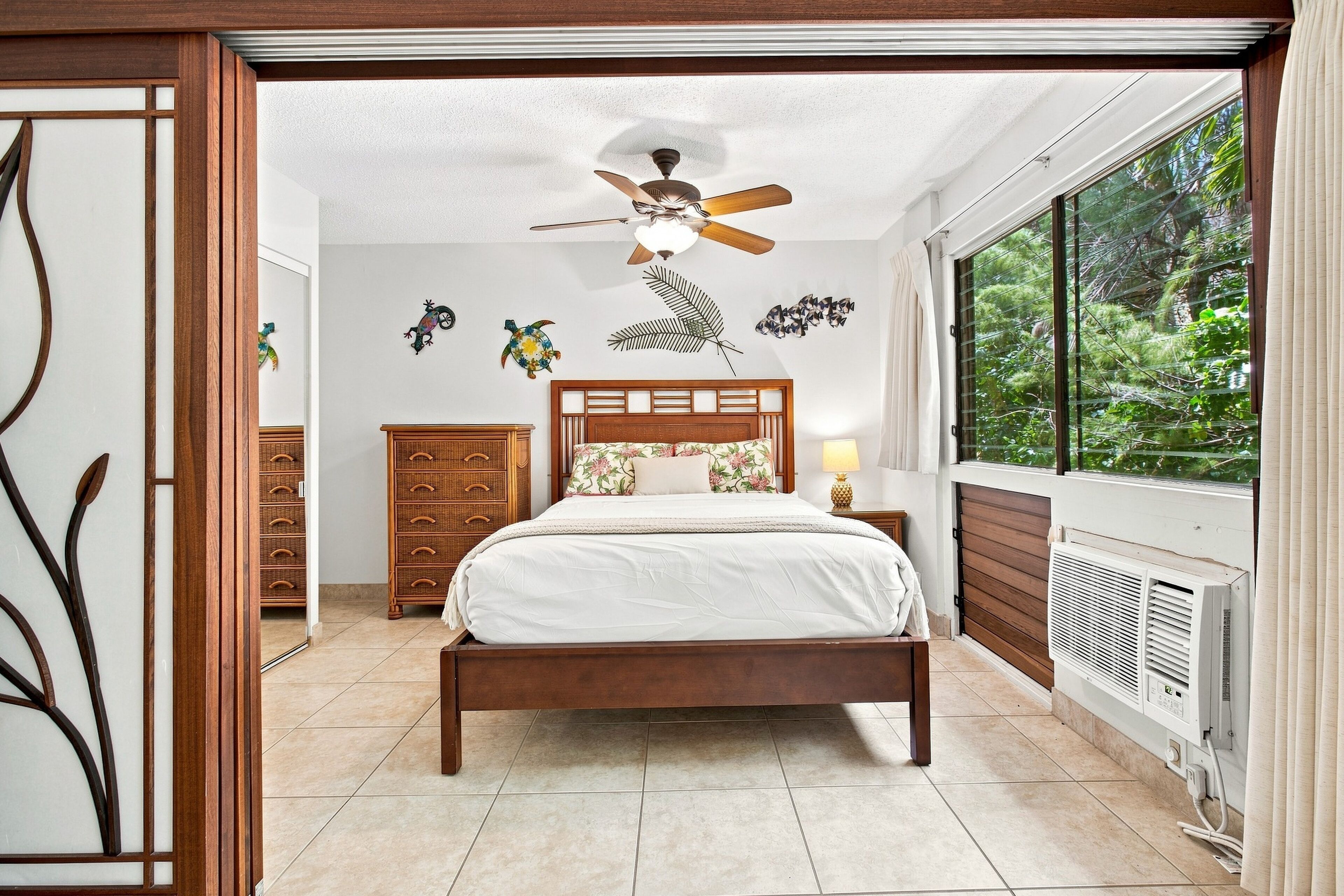 Une chambre avec un lit en bois, un ventilateur de plafond, une fenêtre donnant sur un paysage verdoyant et un mur orné d’œuvres d’art décoratives.