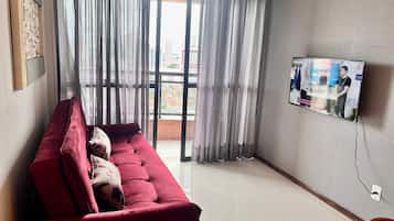 Uma sala de estar com um sofá vermelho, uma TV fixada na parede e vista da cidade através de grandes janelas.