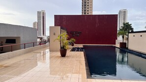 Zona de piscina en la azotea con un muro rojo, plantas en macetas y vistas de edificios altos.