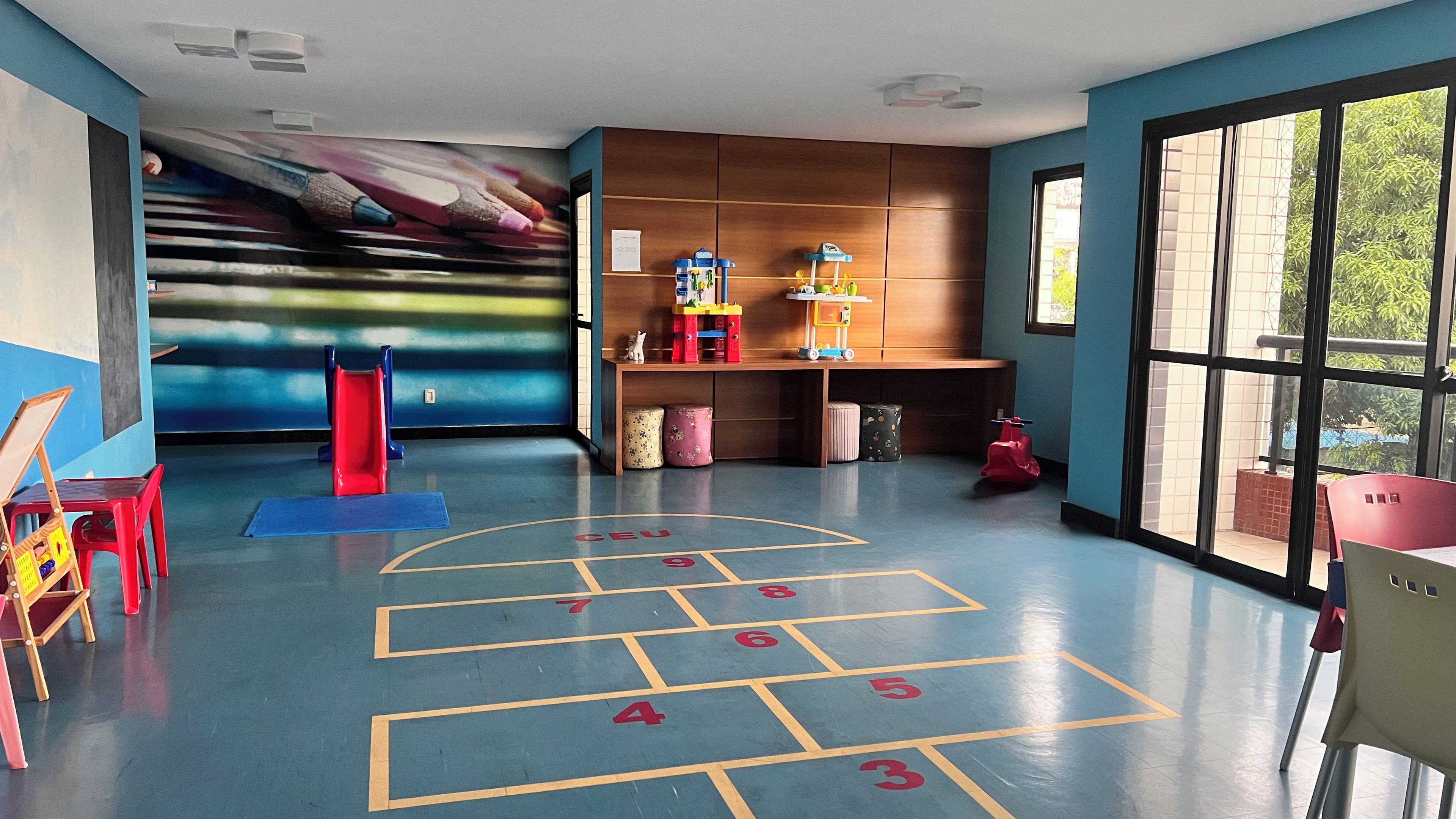 Espaço infantil com um campo de amarelinha, um escorrega e brinquedos nas prateleiras.