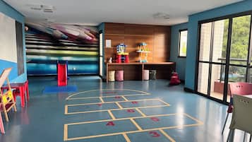 Espaço infantil com um campo de amarelinha, um escorrega e brinquedos nas prateleiras.