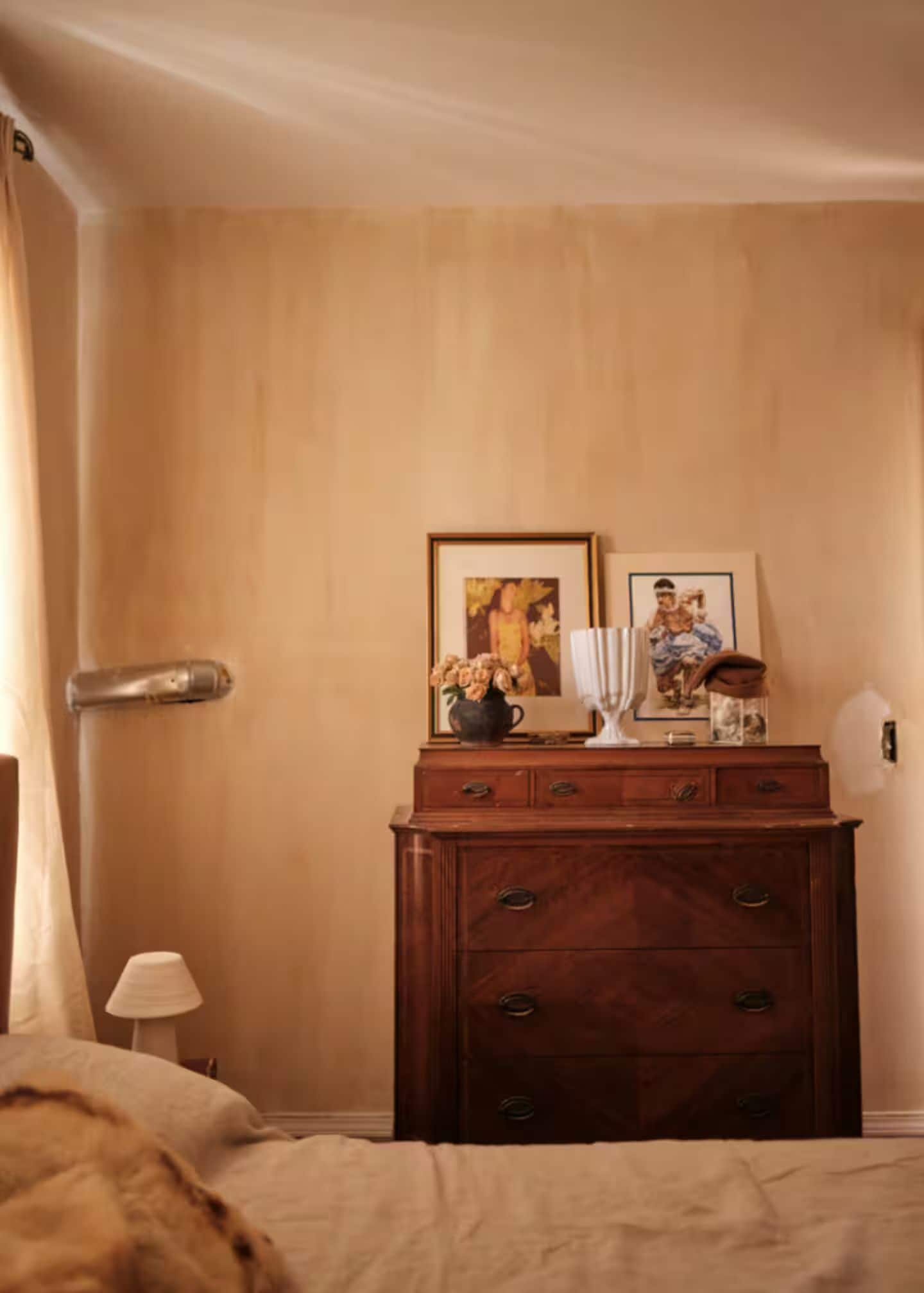A room with a wooden dresser, a bed, a lamp, and two framed pictures on the wall.