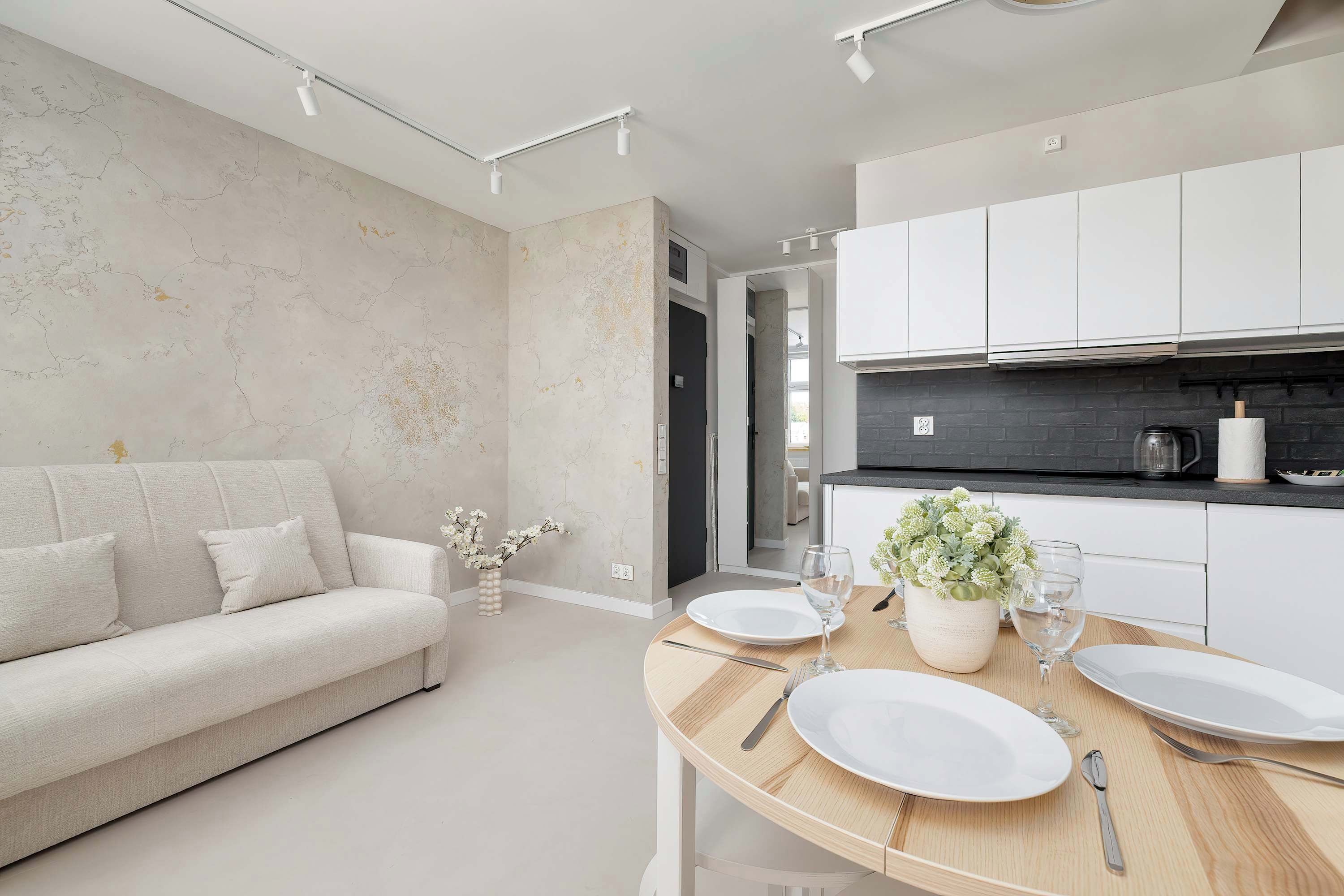 A bright open-plan living area combines a cozy sofa with a wooden dining table set for guests. Modern white cabinetry lines the kitchenette wall, creating a clean and functional living space.