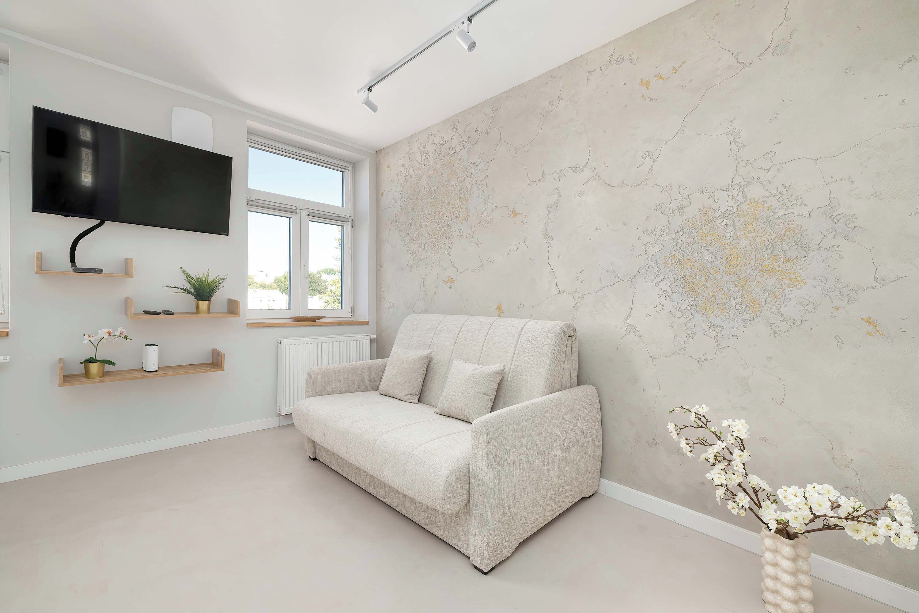 A restful corner provides a plush light-colored sofa facing a wall-mounted television and decorative wooden wall shelves. Soft neutral textures create a warm and relaxing atmosphere.