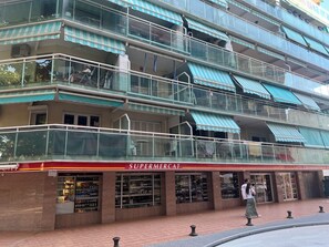 Un immeuble à plusieurs étages abritant un supermarché au rez-de-chaussée, avec des balcons en verre et des auvents à rayures.