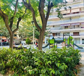 Un edificio residencial de varios pisos con balcones, un parque infantil con juegos verdes, y una calle con coches estacionados.