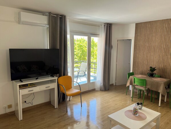 Un salon comprenant un téléviseur, un bureau, une chaise, une table à manger avec des chaises, et donnant sur l’extérieur.