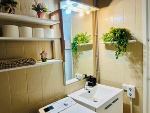 Une salle de bain avec un lavabo blanc, un miroir, des toilettes et des étagères où sont rangés les produits de toilette et des plantes.