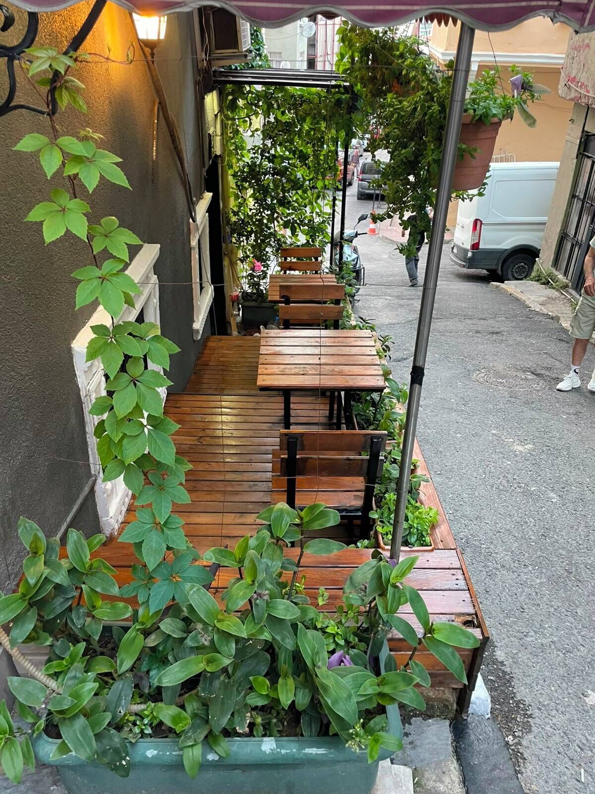An outdoor seating area with wooden tables and chairs, surrounded by potted plants and a metal pole.