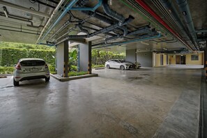 A parking garage with two cars parked under a covered area, concrete floors, and visible structural elements like beams and pipes. - Seashore Beach Residences by NLA (Choeng Thale)