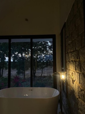 A bathroom with a freestanding bathtub, a stone wall, and a view of trees through a large window. - Gulabbagh jawai  (Jawai Bandh)