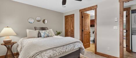 A bedroom with a bed, a nightstand, a lamp, a ceiling fan, and a door leading to a bathroom.