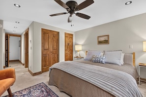A bedroom with a bed, a ceiling fan, wooden doors, a nightstand, a lamp, and a painting on the wall. - Beaver Village Unit 624 2 bed remodeled, private deck w/forest views (Winter Park)