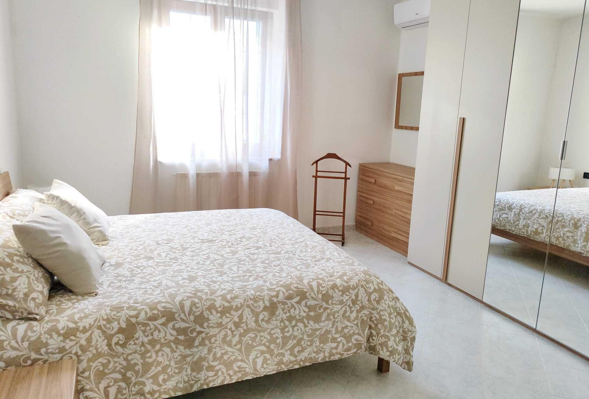 Ein Schlafzimmer mit einem Bett, einem Schminktisch, einem Spiegel und einem Fenster mit durchsichtigen Vorhängen.