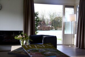 A living room with a sofa, a magazine, and a view through a glass door to an outdoor area with plants. - Bungalow in Hoge Hexel with swimming pool (Hoge Hexel)