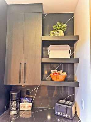 A kitchen with dark wooden cabinets, a marble countertop, a coffee maker, a toaster, and a shelf with a plant and a basket of snacks.