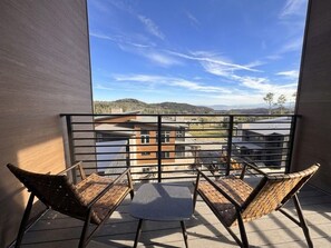 En balkon med to fletstole og et lille bord, med udsigt over et boligområde med huse og bakker.
