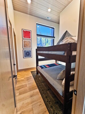 Een stapelbed met een geruit sprei, een raam met uitzicht op bomen, en lijstwerken aan de muur.